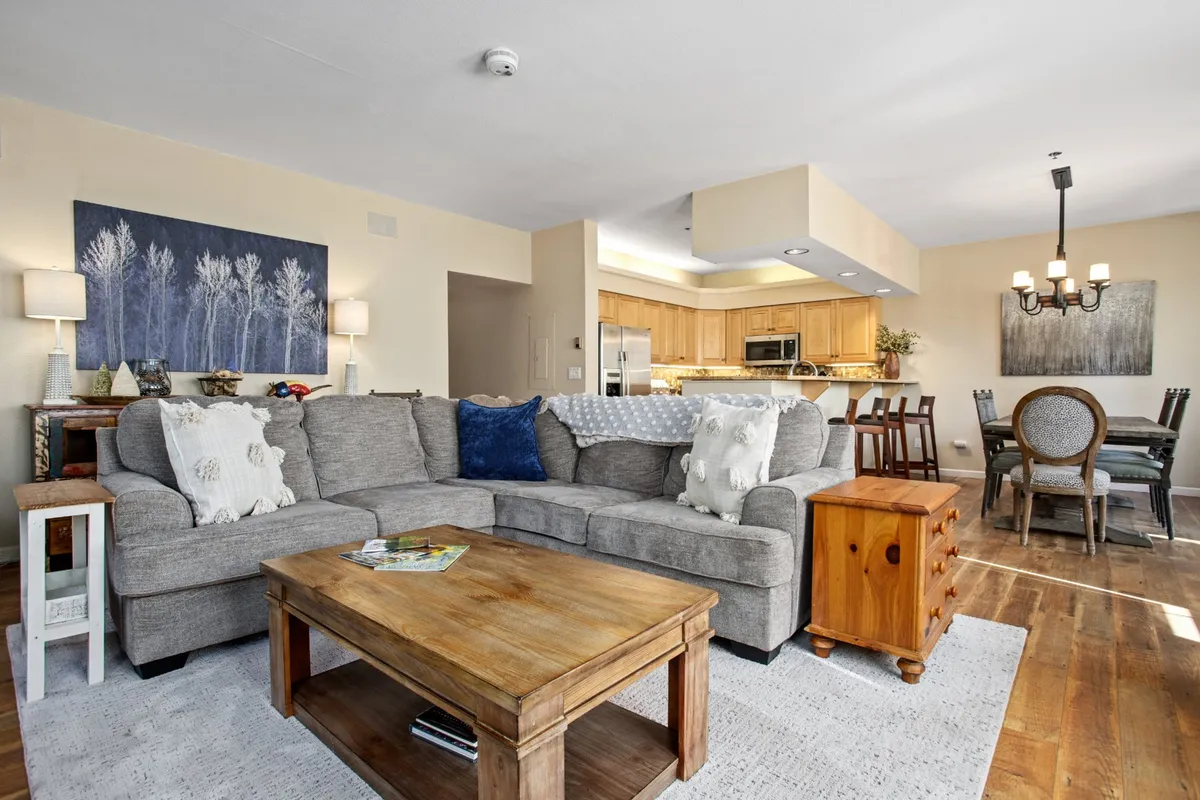 A welcoming open-concept living and dining space featuring a large gray sectional sofa, a wooden coffee table, artwork on the wall, and a dining table with chairs, all bathed in natural light. The arrangement suggests ample space for relaxing, entertaining, or gathering for meals.
