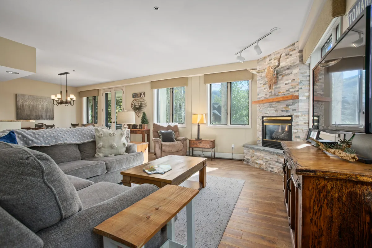 The image showcases a cozy living room in a Steamboat Springs condo, featuring a plush sofa, inviting stone fireplace, spacious wooden coffee table, and expansive windows that fill the space with natural light and frame views of the surrounding nature.