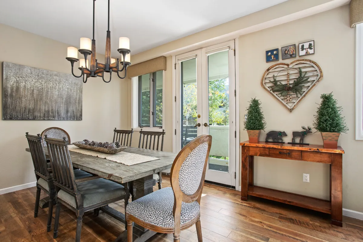 A cozy dining area with a wooden table set beneath a chandelier, small potted plants adding a homely touch, and double doors that open out to an outdoor space. Heart-shaped wall art and the proximity to the exterior reinforce the warmth and connection to the outdoors.