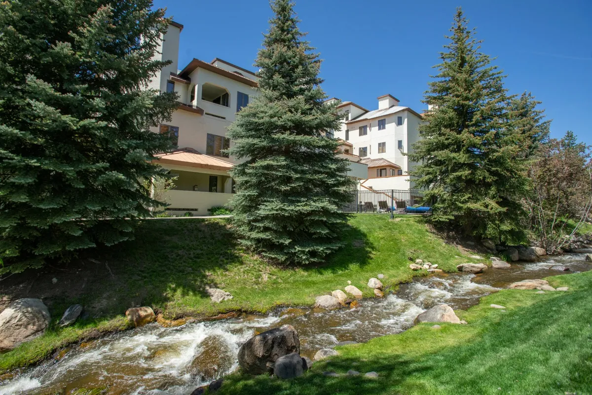 This exterior shot captures Burgess Creek flowing past tall evergreens, with the multi-story building overlooking the serene scene. The natural setting highlights the unique creekside location and the peaceful retreat atmosphere offered to guests.