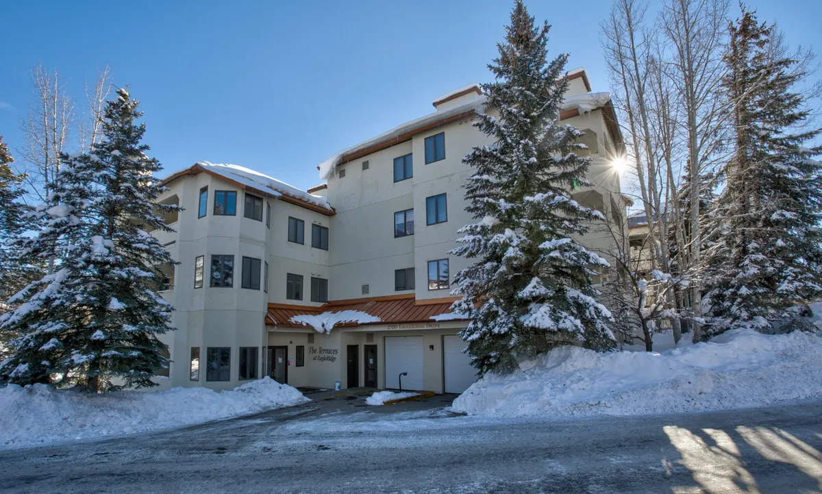 A winter exterior shot highlighting modern architecture set against a backdrop of snow-covered trees and a blue sky. Sunlight glints off the building's windows, emphasizing a welcoming and vibrant winter environment, perfectly evocative of a mountain resort atmosphere.
