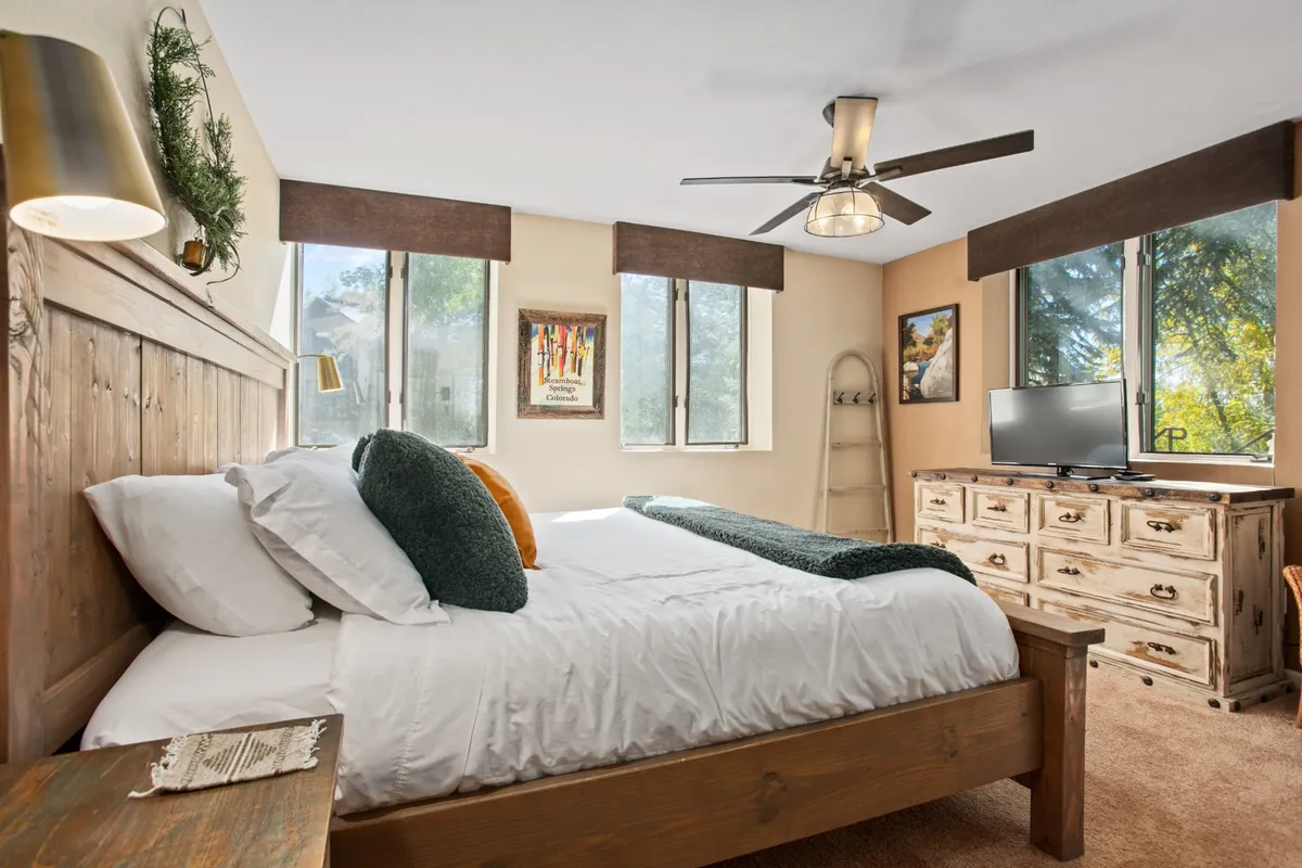 A serene bedroom featuring a wooden bed frame dressed in crisp white linens and decorative pillows, with a TV atop a dresser, artwork on the wall, and large windows allowing abundant natural light. The space feels both comfortable and private, ideal for restful moments.