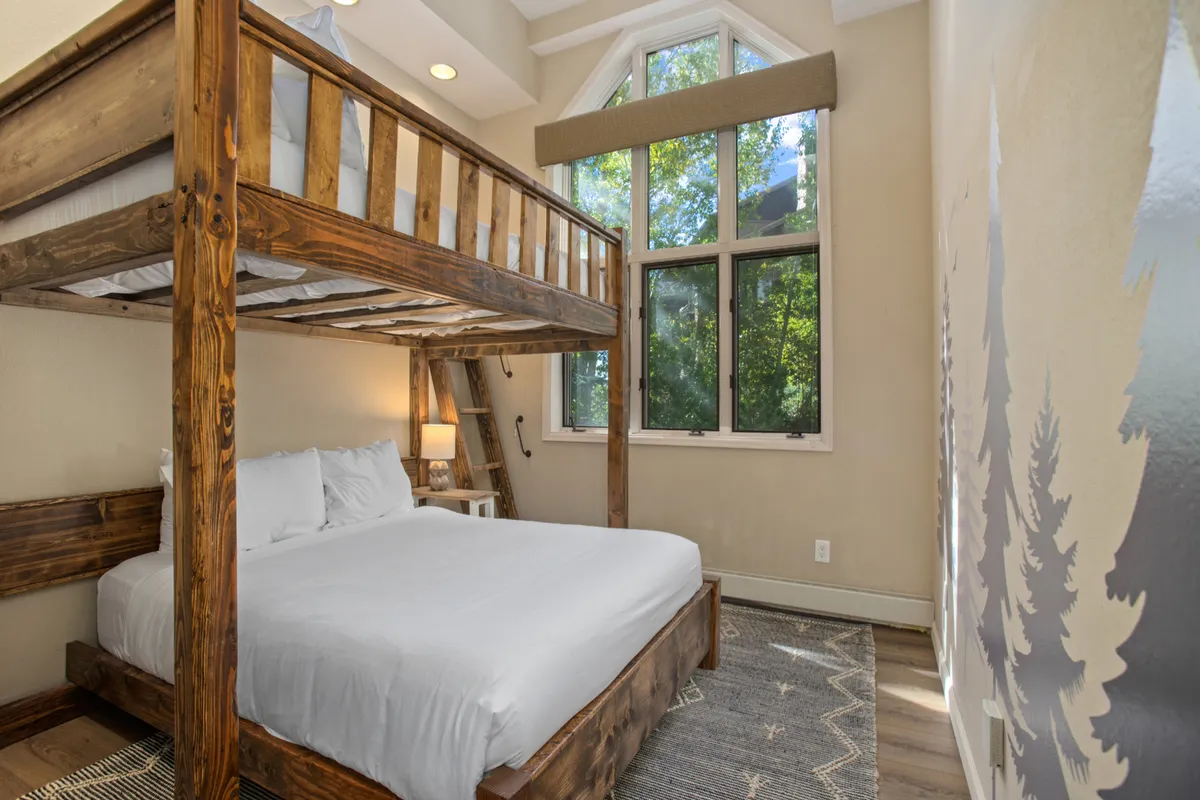 A bright bunk room with a sturdy bunk bed (full-over-queen), large windows letting in plenty of daylight, wooden touches, and a cozy rug. The setting feels playful and ideal for kids or groups, offering flexibility and a welcoming vibe.