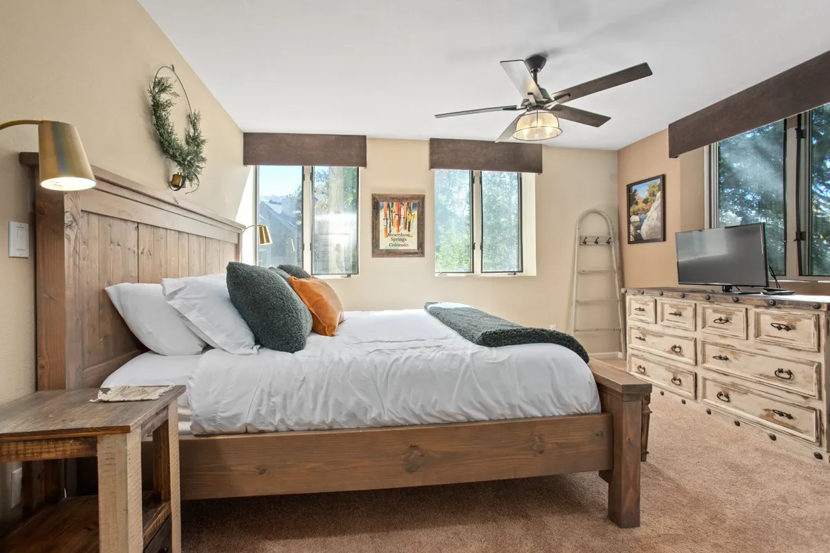 A welcoming bedroom with a solid wooden bed, decorative pillows, a dresser, and generous windows. Soft light fills the room, creating a cozy spot for reading or relaxing at any time of day.